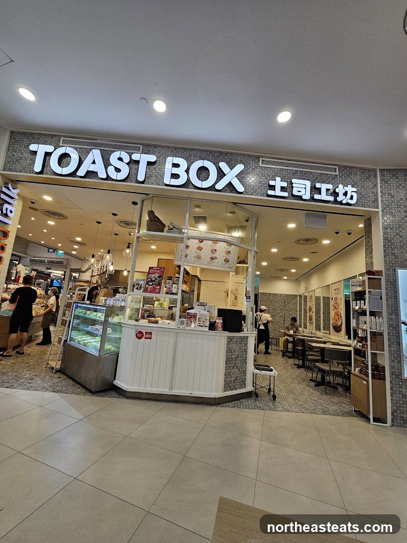 The exterior of a Toast Box cafe with its prominent signage. Inside, display cases show food items, and seating areas are visible.