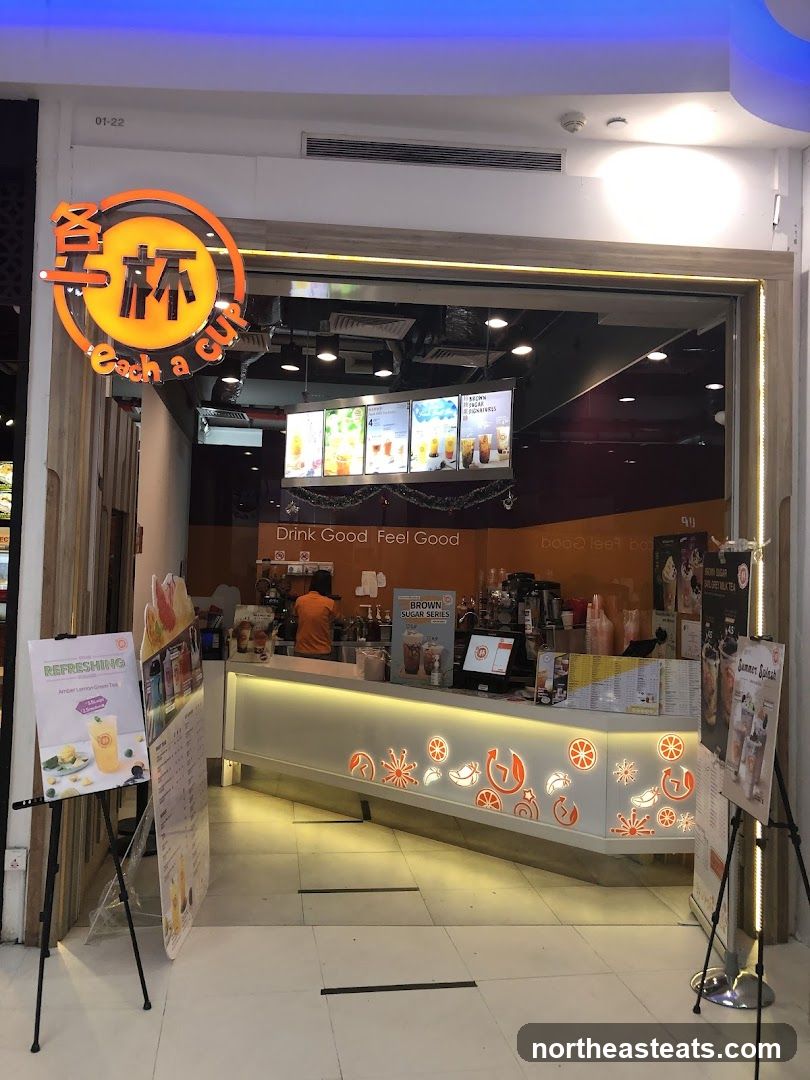 The exterior of a bubble tea shop with a prominent orange and yellow logo that says "Each a cup". Inside, a counter is visible with drinks displayed on screens above. The shop has a modern design with orange and white accents.