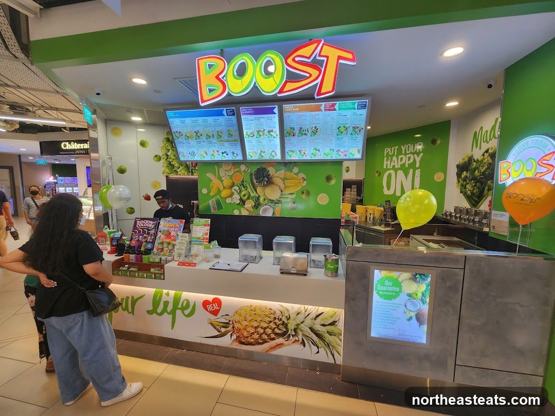 A Boost Juice Bar storefront with a prominent "BOOST" logo. The counter displays various juices and promotional materials. Two people are visible at the counter. Balloons in yellow and orange are placed around the store.