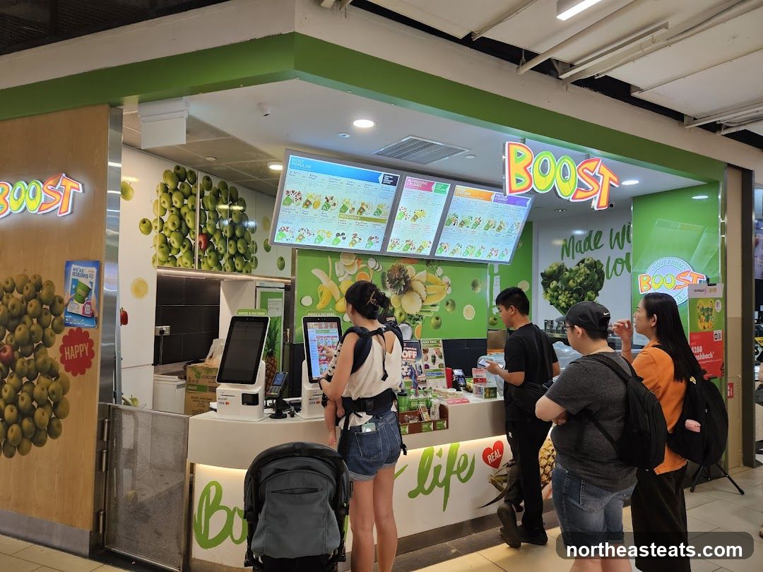 A Boost Juice Bar storefront with a prominent "BOOST" logo above the counter. The interior features digital menus displaying various drinks and food items, along with fruit-themed decor. Customers are seen ordering and waiting.