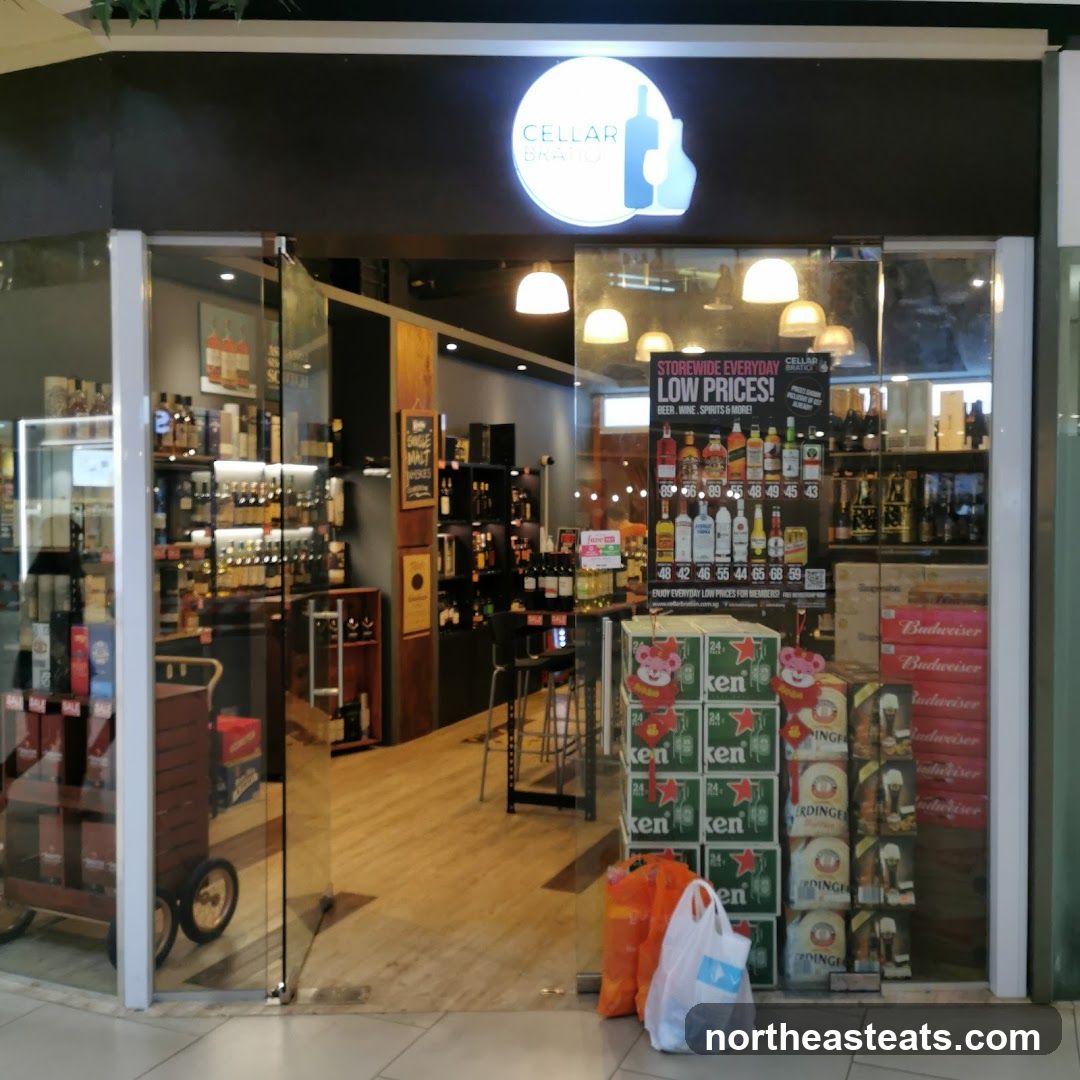 The exterior of a liquor store with a large glass storefront. Inside, shelves are stocked with bottles of wine and spirits. A promotional poster displays "STOREWIDE EVERYDAY LOW PRICES!" for beer, wine, spirits, and more, with prices listed for various bottles. Cases of beer and bottles of Budweiser are stacked near the entrance.