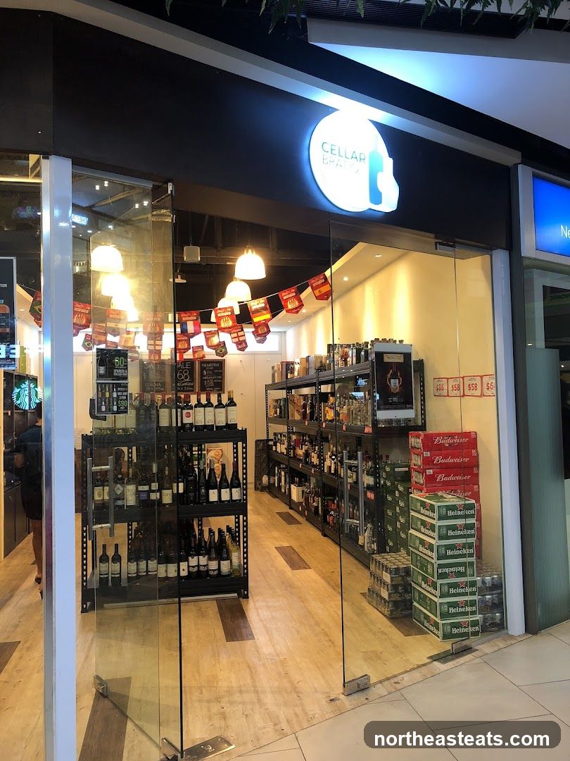 The exterior of a wine and liquor store with a prominent illuminated sign that reads "CELLAR BRATO". The storefront features large glass doors and windows, revealing shelves stocked with bottles of wine and spirits. Stacks of beer cases, including Budweiser and Heineken, are visible near the entrance. Decorative flags hang inside the store.