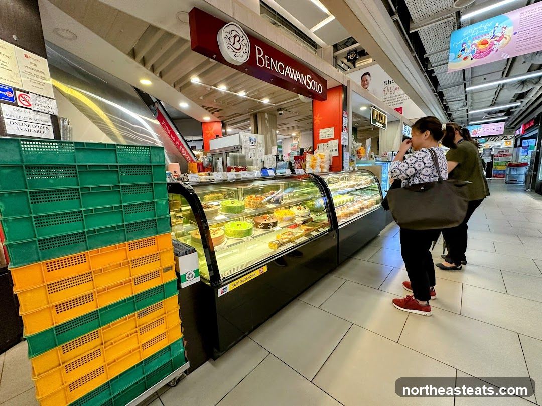 A display case filled with cakes and pastries is visible in front of a shop. Two women are standing in line to order. Stacks of green and yellow crates are to the left of the display.