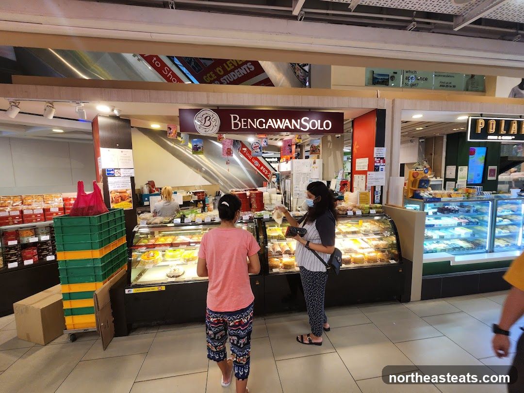 A view of the Bengawan Solo shop front with display cases filled with pastries and cakes. Two customers are visible, one interacting with a staff member behind the counter. Stacks of green and yellow crates are to the left of the shop.