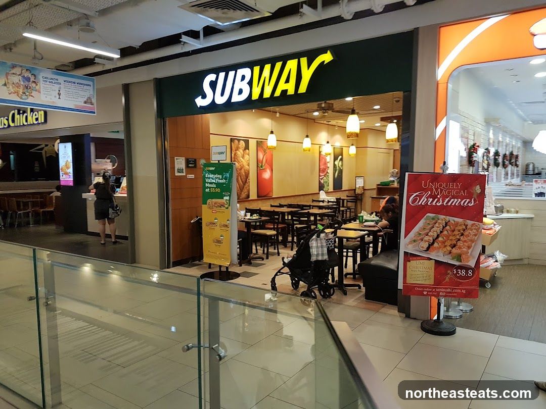The exterior of a Subway restaurant with its prominent green and yellow signage. Inside, tables and chairs are visible, along with food displays. A promotional sign for a Christmas sushi platter is placed outside the entrance.