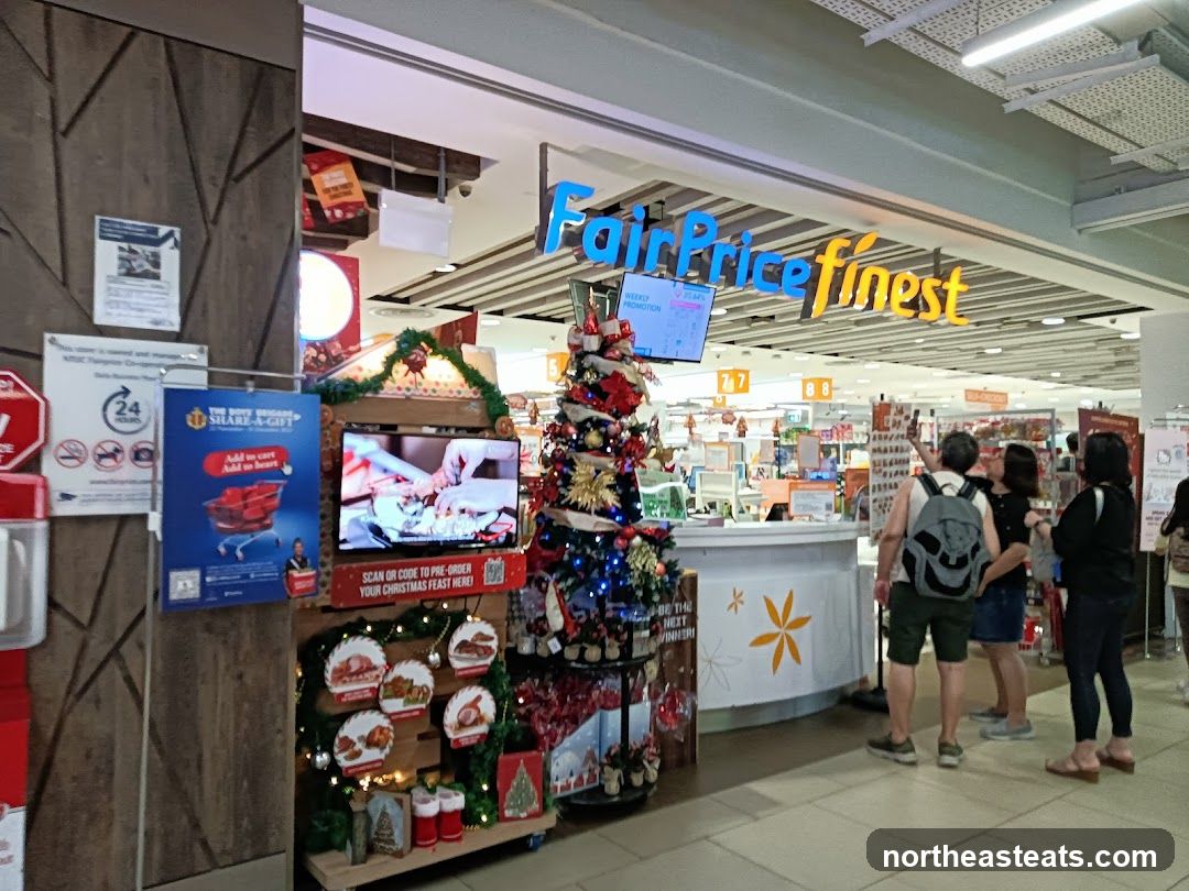 The exterior of a FairPrice Finest store with a Christmas theme. A large sign with the store's name is visible above the entrance. In front of the store, there is a decorated Christmas tree, a display screen showing a cooking demonstration, and various promotional materials related to Christmas feasts. Several customers are standing near the entrance.