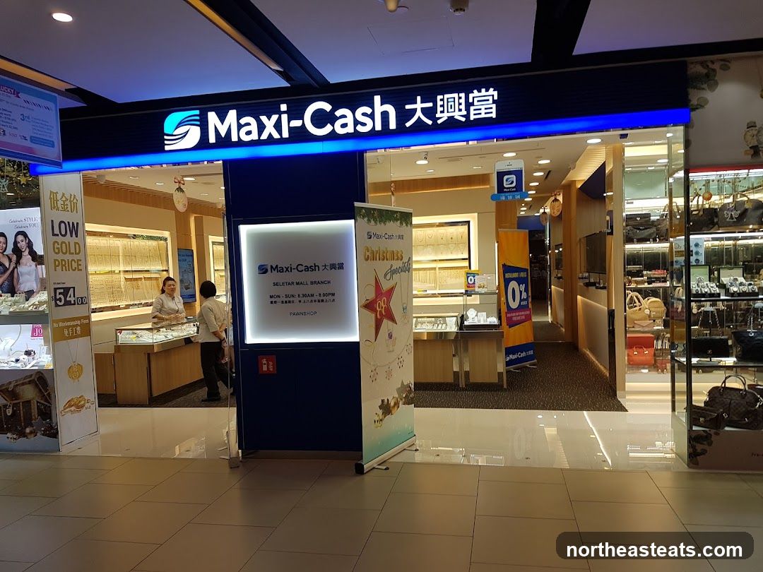 The exterior of a Maxi-Cash pawnshop branch. The prominent blue and white signage with the Maxi-Cash logo is visible above the entrance. Inside, display cases filled with jewelry are visible, along with staff members attending to customers. Promotional posters and banners are placed outside the shop, advertising low gold prices and a 0% interest offer.