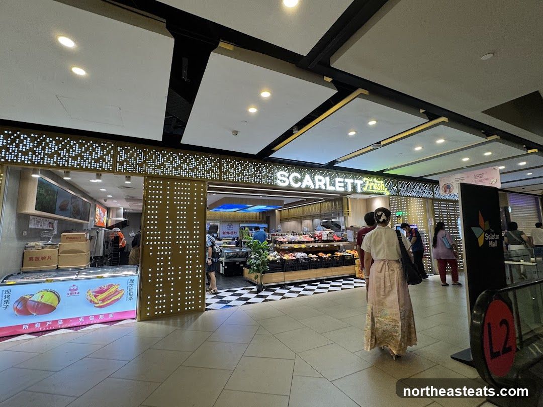 A view of the Scarlett Fresh storefront in a mall. The store has a modern facade with illuminated decorative panels. Inside, shelves are stocked with produce and other goods. A sign for Seletar Mall is visible to the right.