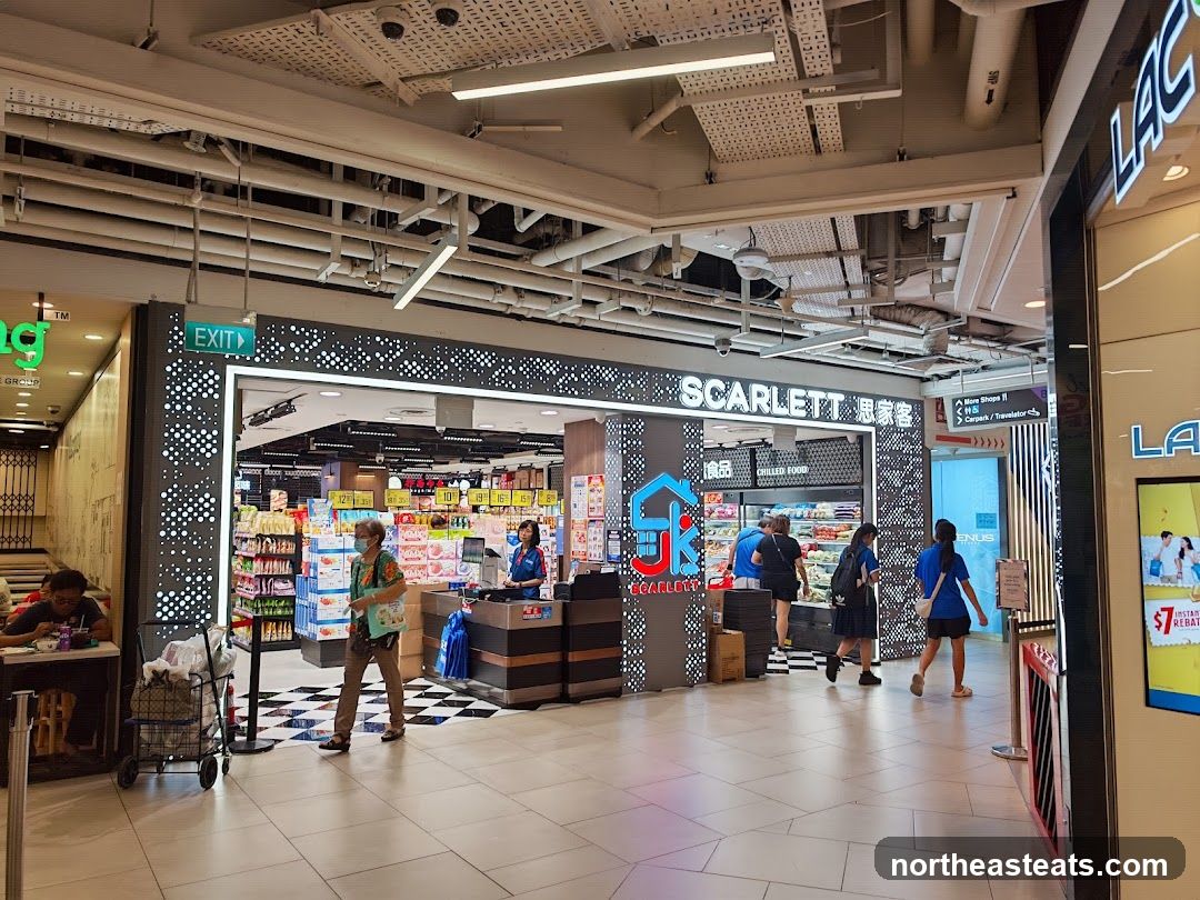 The entrance to a supermarket named Scarlett, with its logo and name displayed prominently above the entrance. The store appears to be well-stocked with various products visible inside, including chilled food. Shoppers are seen entering and exiting the store.
