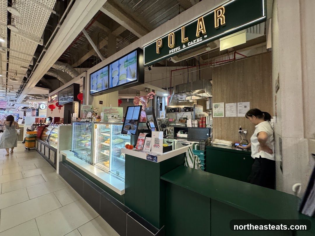 A view of the Polar Puffs & Cakes storefront, showcasing a display case filled with pastries and cakes. A staff member is visible behind the counter. The shop's signage is prominently displayed above the counter.