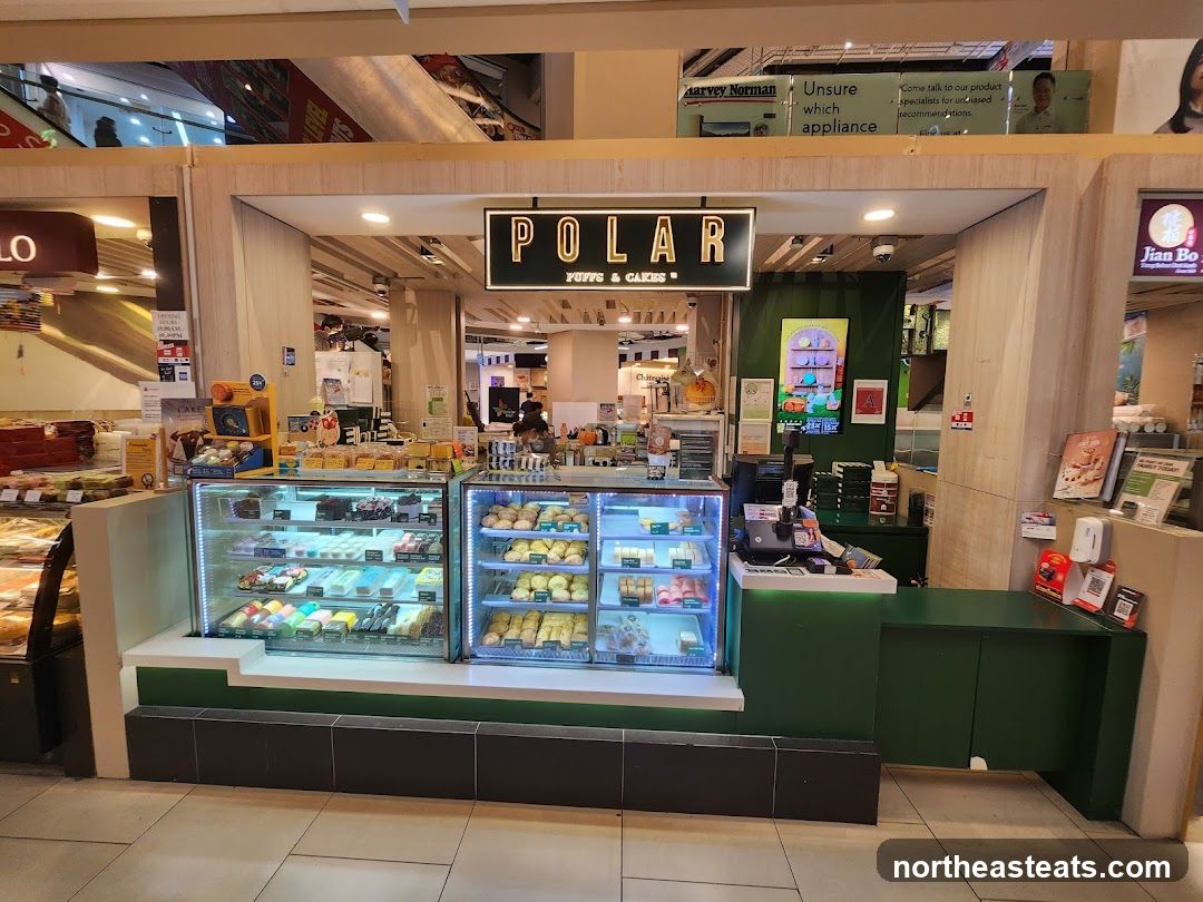 A brightly lit bakery counter displaying a variety of colorful cakes and pastries in refrigerated display cases. The shop's name, "POLAR PUFFS & CAKES," is prominently displayed above the counter. The interior of the mall is visible in the background, with other shops and escalators.