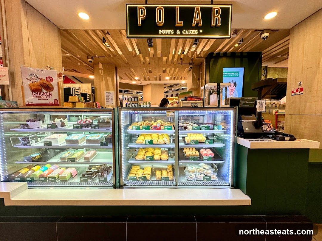 A display of various cakes and pastries in refrigerated display cases at Polar Puffs & Cakes. The shop's name is visible on a sign above the display.