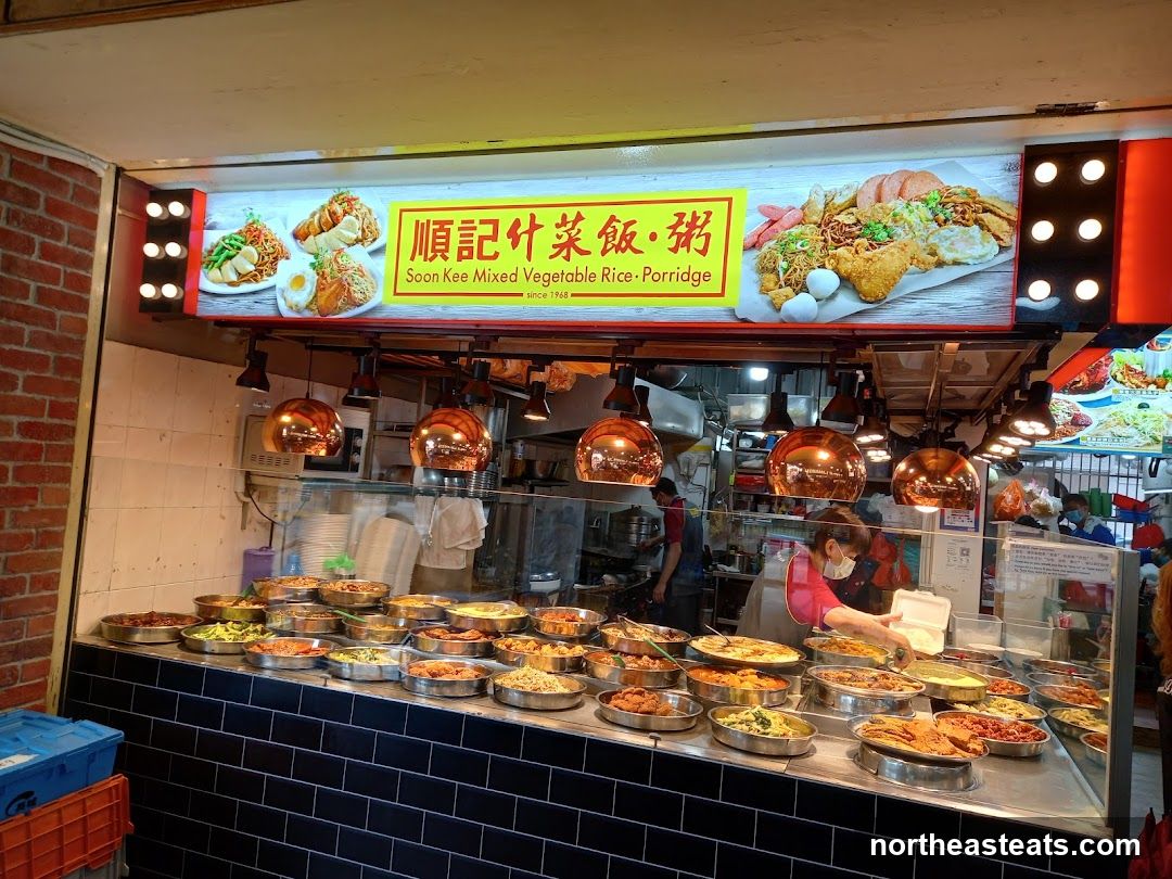 A food stall with a wide variety of dishes displayed in metal trays. The stall has a large illuminated sign with images of food and the name of the stall in English and Chinese. The sign reads "Soon Kee Mixed Vegetable Rice • Porridge Since 1968". There are several employees visible behind the counter, serving customers. The stall appears to be busy.