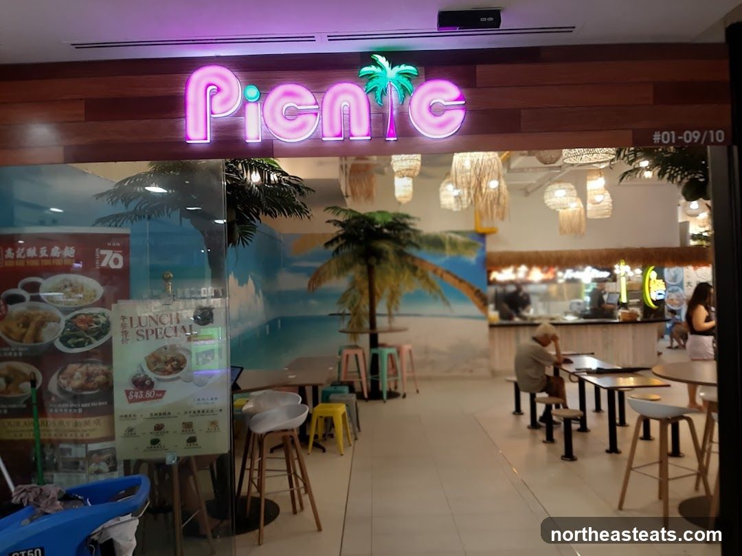 The storefront of a restaurant called Picnic, with neon pink lettering and a palm tree logo. The entrance has large glass windows displaying menus and food items. Inside, there are tables and chairs, and a counter area with staff. The decor includes tropical-themed murals and woven light fixtures.