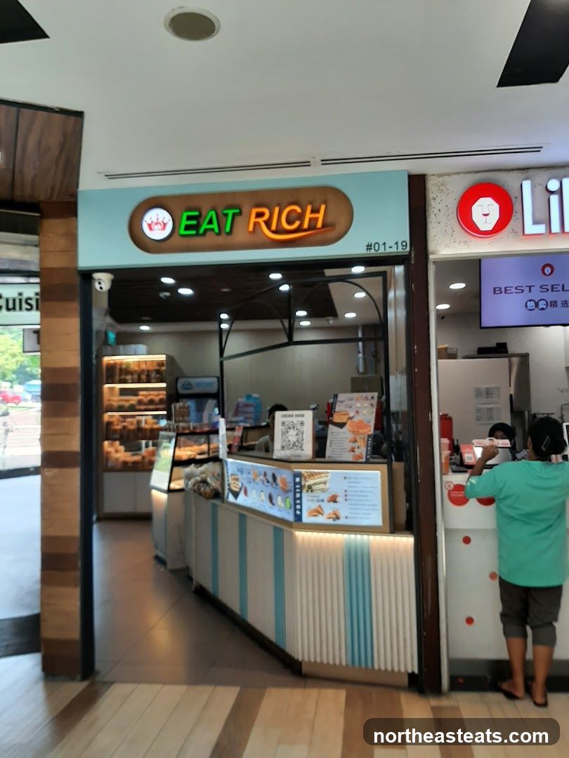 A brightly lit shop front with a prominent neon sign that reads "EAT RICH". Below the sign, a counter displays various food items and a menu board. The shop appears to be a food stall or small eatery within a larger establishment.