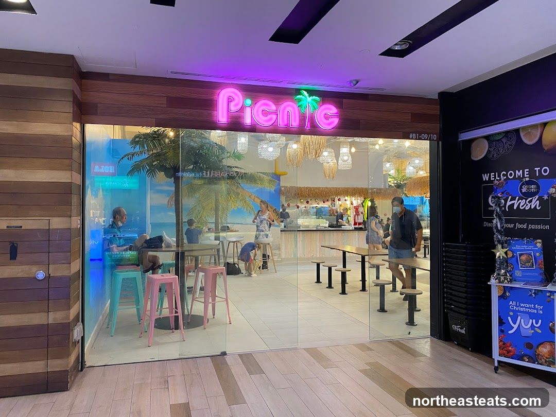 The exterior of a restaurant called Picnic, with neon pink signage and a tropical mural visible through the glass. Inside, tables and chairs are set up, and people are dining. A promotional stand with Christmas-themed decorations is to the right.