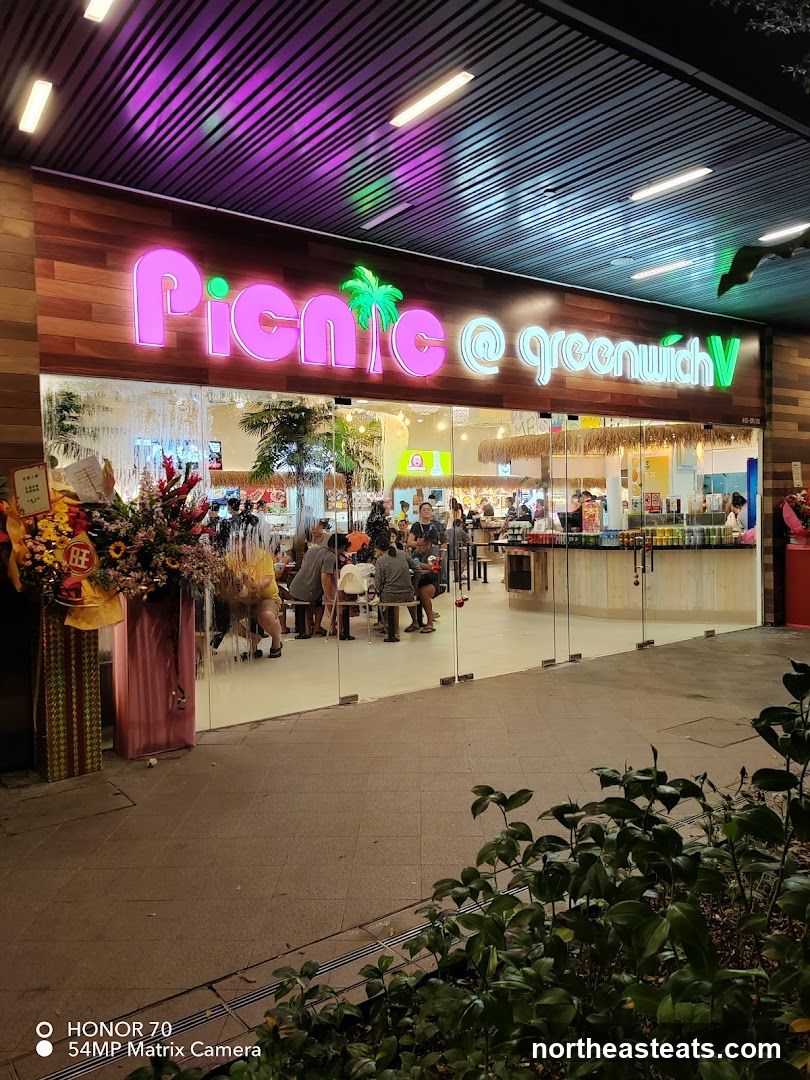 The exterior of a restaurant with a large neon sign that reads "Picnic @ greenwichV". The restaurant has large glass doors and windows, and inside, people are seated at tables. There are tropical plants and thatched roof elements visible inside, suggesting a relaxed, outdoor-themed dining experience. Floral arrangements are placed outside the entrance.