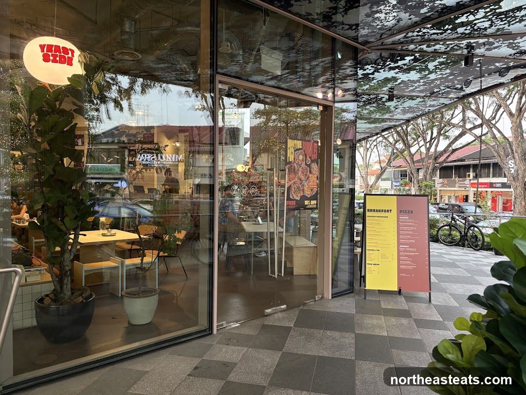 The exterior of a modern cafe with large glass windows and doors. A prominent round light fixture with the words "YEAST SIDE" in red letters hangs inside. Inside, tables and chairs are visible, along with plants. Outside, a menu board displays breakfast and pizza options. A bicycle is parked nearby. The cafe is situated on a street with other businesses and trees lining the sidewalk.