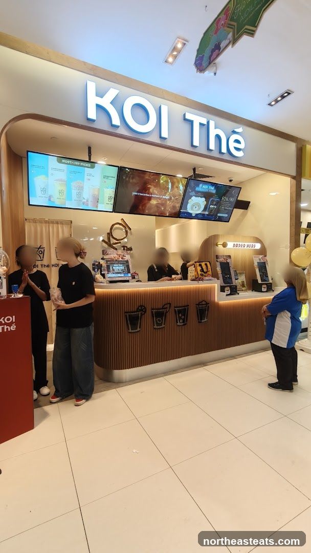 A brightly lit KOI Thé bubble tea shop interior with a prominent "KOI Thé" sign. Digital screens display menu items and promotions. Staff members are visible behind the counter, and customers are interacting with the ordering system. The counter has a wooden slatted design with stylized drink icons.