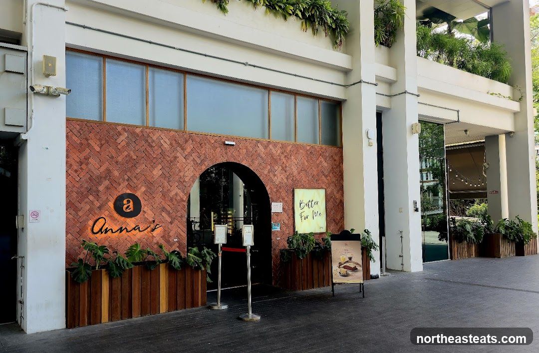 The exterior of a restaurant with a brick facade and an arched entrance. The restaurant's name, "Anna's", is illuminated in orange script above a black circular logo with a white 'a'. Planters with lush green plants line the base of the facade. To the right of the entrance, a sign with the text "Better For Me" is displayed. An A-frame sign in front of the entrance advertises a dessert item.