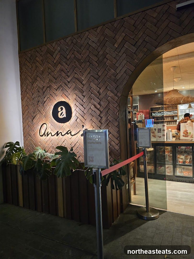 The exterior of a shop with a brick herringbone pattern wall. A sign with a stylized 'a' and the name 'Anna's' is illuminated. Plants are arranged in front of the lower wooden paneling. A queue barrier with a red ribbon leads towards the entrance, where a sign reads 'Our queue starts here.' Inside, a counter with display refrigerators and shelves is visible.