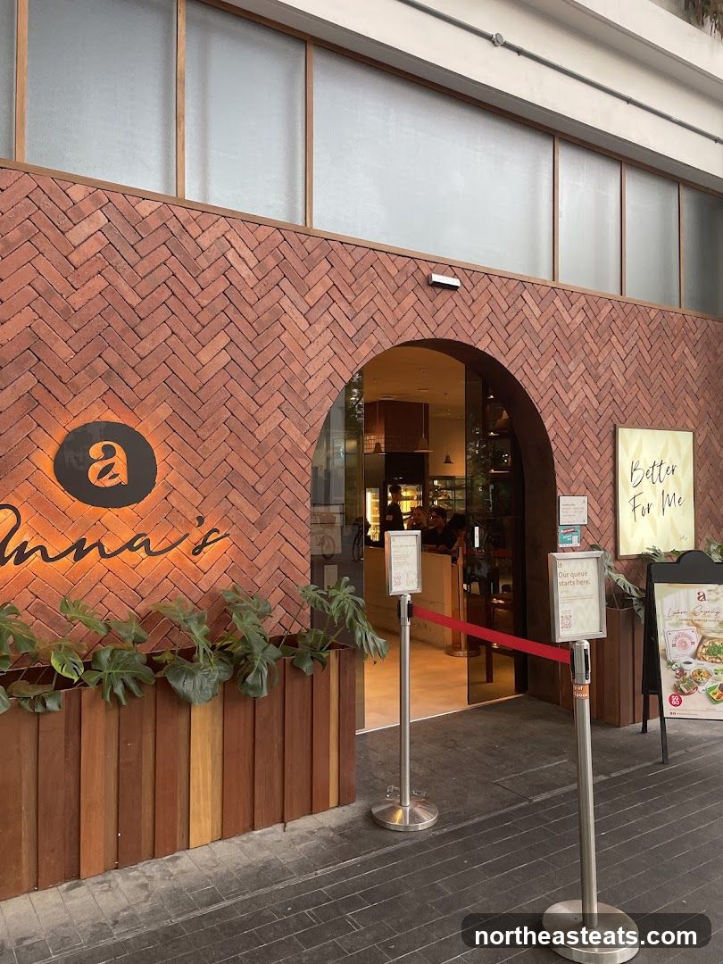 The exterior of a bakery with a herringbone brick facade. An arched entrance leads into the shop, where a counter and seating are visible. A large illuminated sign with the letter 'a' and the word 'Anna's' is mounted on the brick wall. Planters with lush green plants are situated in front of the entrance. Queue barriers with red tape are set up outside the doorway. A sign on an easel displays food items and promotions.