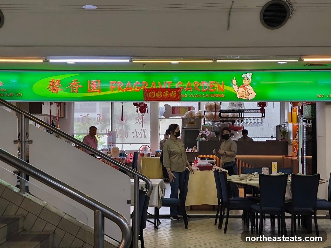 A restaurant storefront with a large green sign displaying the name "FRAGRANT GARDEN" in English and Chinese characters. Below the name, it reads "NG YUAN CATERING". A cartoon chef giving a thumbs up is also on the sign. The interior of the restaurant is visible through the windows, showing tables, chairs, and staff.