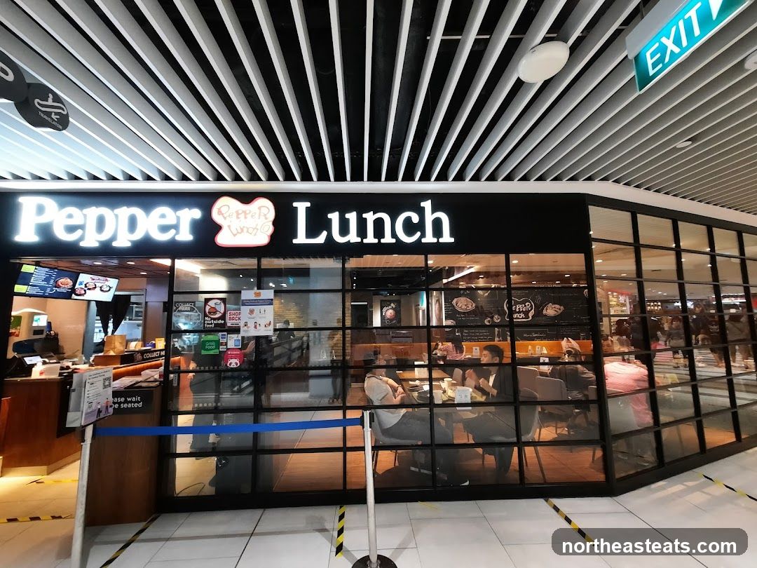 The exterior of a Pepper Lunch restaurant with large glass windows and a prominent sign. Inside, customers are seated at tables. An exit sign is visible above the entrance.