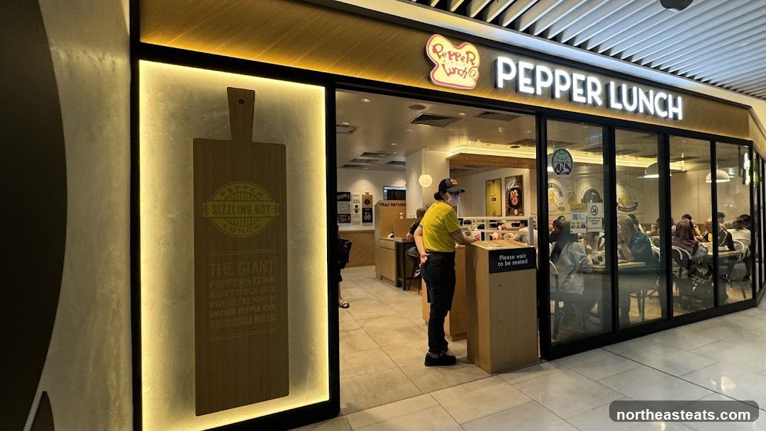 The exterior of a Pepper Lunch restaurant with large glass windows and a prominent sign. Inside, customers are seated at tables, and a staff member in a yellow shirt stands near the entrance. A wooden board with menu items is displayed on the left side of the entrance.