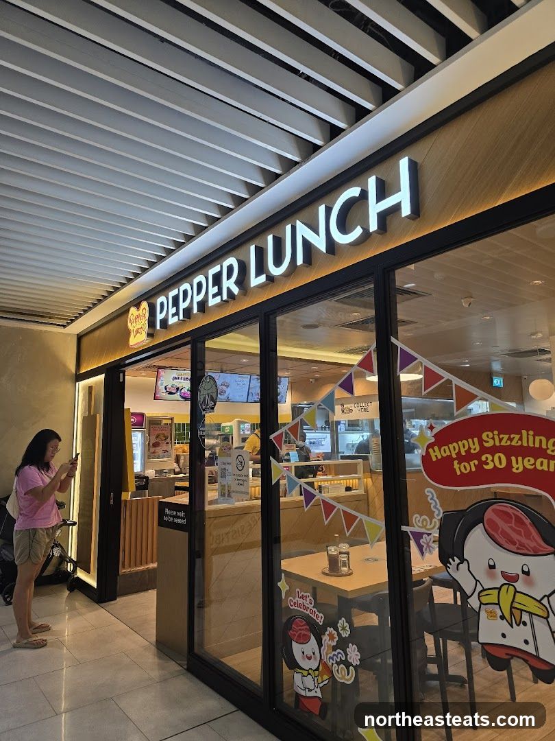 A woman stands outside a Pepper Lunch restaurant, looking at her phone. The storefront features a large illuminated sign that reads "PEPPER LUNCH" and a logo. The windows display colorful decorations and cartoon characters, with one sign saying "Happy Sizzling for 30 years" and another "Let's Celebrate!". Inside, tables and chairs are visible, along with a counter area.