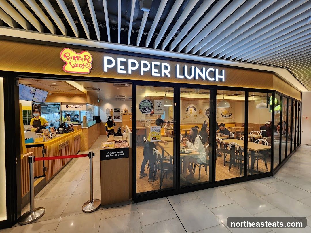 The exterior of a Pepper Lunch restaurant with large glass windows and a prominent sign displaying the restaurant's name. Inside, customers are seated at tables, and staff are visible behind the counter.