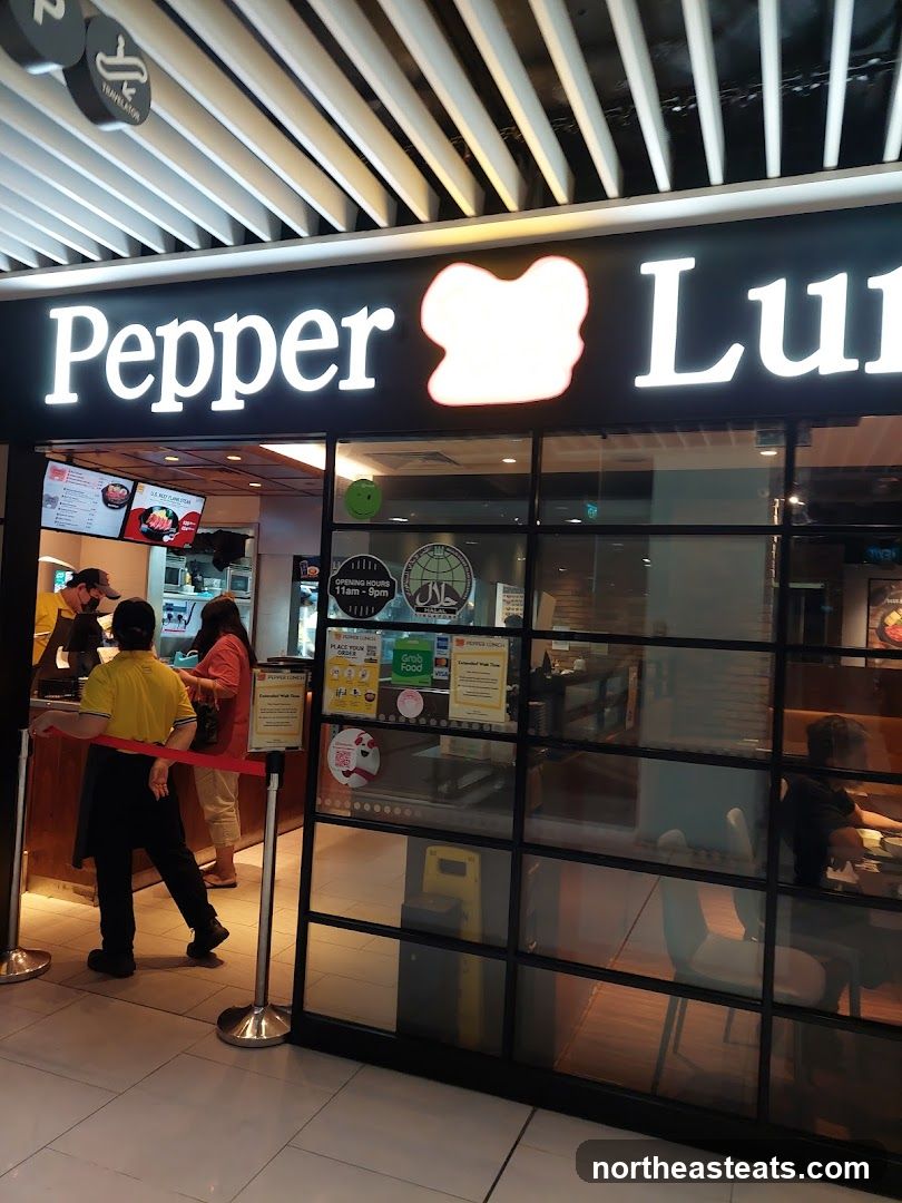 The exterior of a Pepper Lunch restaurant with its prominent signage. Inside, staff members are visible behind the counter, and customers are seen queuing. The storefront features large glass windows, allowing a view into the dining area where patrons are seated.