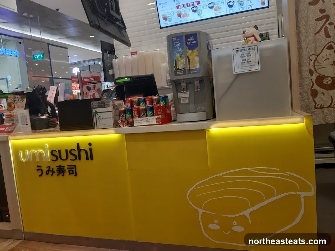 The exterior of a bright yellow Umi Sushi shop with the store name prominently displayed in white and black lettering. A cartoon drawing of sushi is on the lower part of the facade. Behind the counter, there are drinks, a beverage dispenser, and a menu board visible.