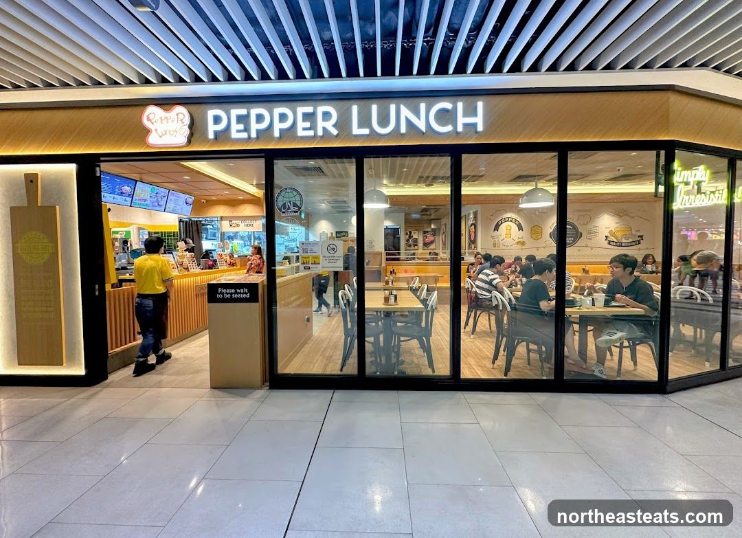 The exterior of a Pepper Lunch restaurant with large glass windows and doors. The restaurant's name is prominently displayed in illuminated white letters above the entrance. Inside, tables and chairs are visible, with customers dining. A counter area with staff serving food is also seen. The overall ambiance appears to be a casual dining establishment.