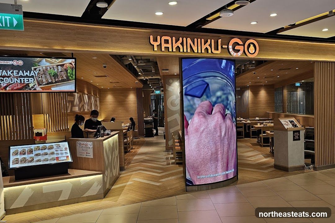 The exterior of a YAKINIKU-GO restaurant. A large digital screen displays a close-up of a hand. There is a takeaway counter with staff members. The restaurant interior is visible with tables and chairs.