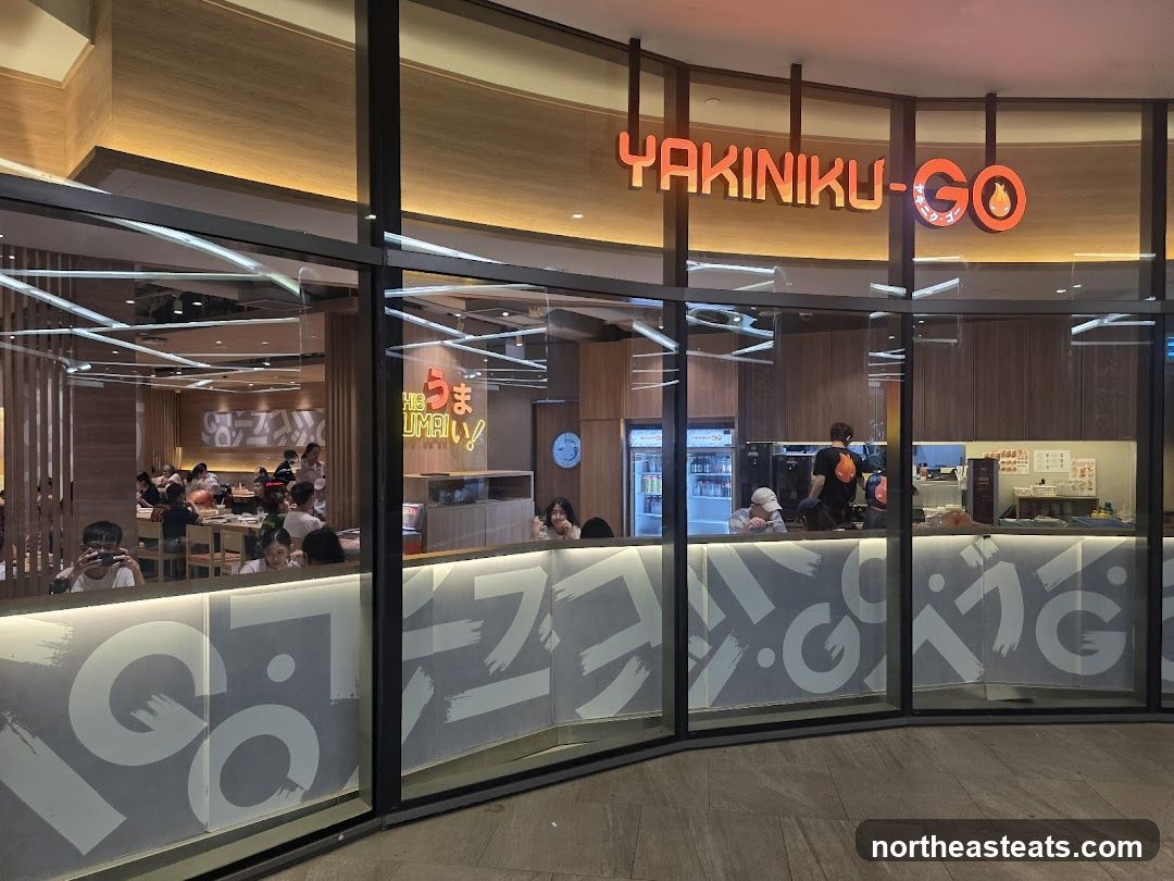 The exterior of a Yakiniku-Go restaurant is visible through large glass windows. The restaurant's name is prominently displayed in orange neon letters above the entrance. Inside, diners are seated at tables, and staff can be seen behind a counter. The interior features wooden paneling and modern lighting. The lower part of the windows has a frosted glass design with abstract white brush strokes and Japanese characters.