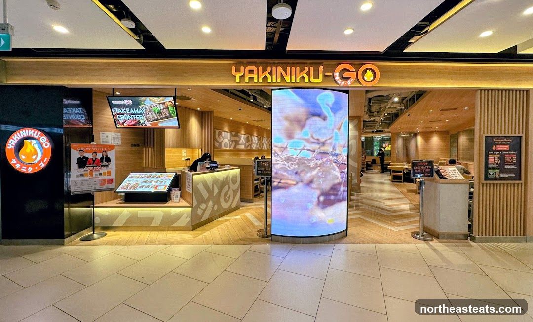 The exterior of a Yakiniku-Go restaurant. The entrance features a prominent sign with the restaurant's name and logo. To the left, a circular logo with flames and Japanese characters is visible. A takeaway counter is advertised on a screen above the counter. A digital display screen is also present at the entrance.