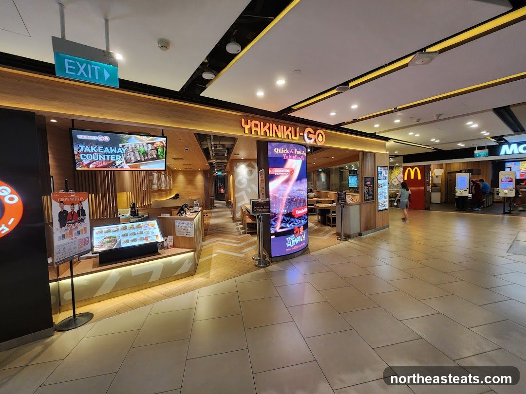 The exterior of a Yakiniku-Go restaurant with a takeaway counter. A digital screen displays "TAKEAWAY COUNTER" and images of grilled food. A menu board with pictures of dishes is also visible. The restaurant has a modern interior with wooden accents. In the background, a McDonald's outlet is visible.