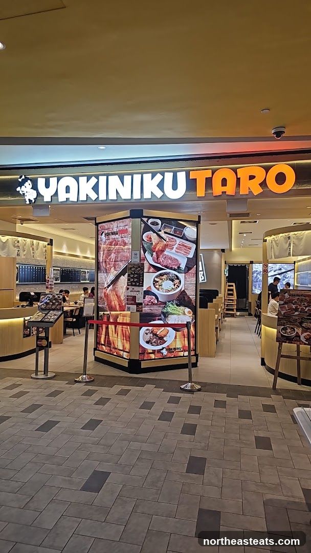 The exterior of a Yakiniku Taro restaurant. The prominent signage above the entrance reads "YAKINIKU TARO" in large orange and white letters. The entrance area features a display with images of various Japanese barbecue dishes. Inside, diners can be seen seated at tables.