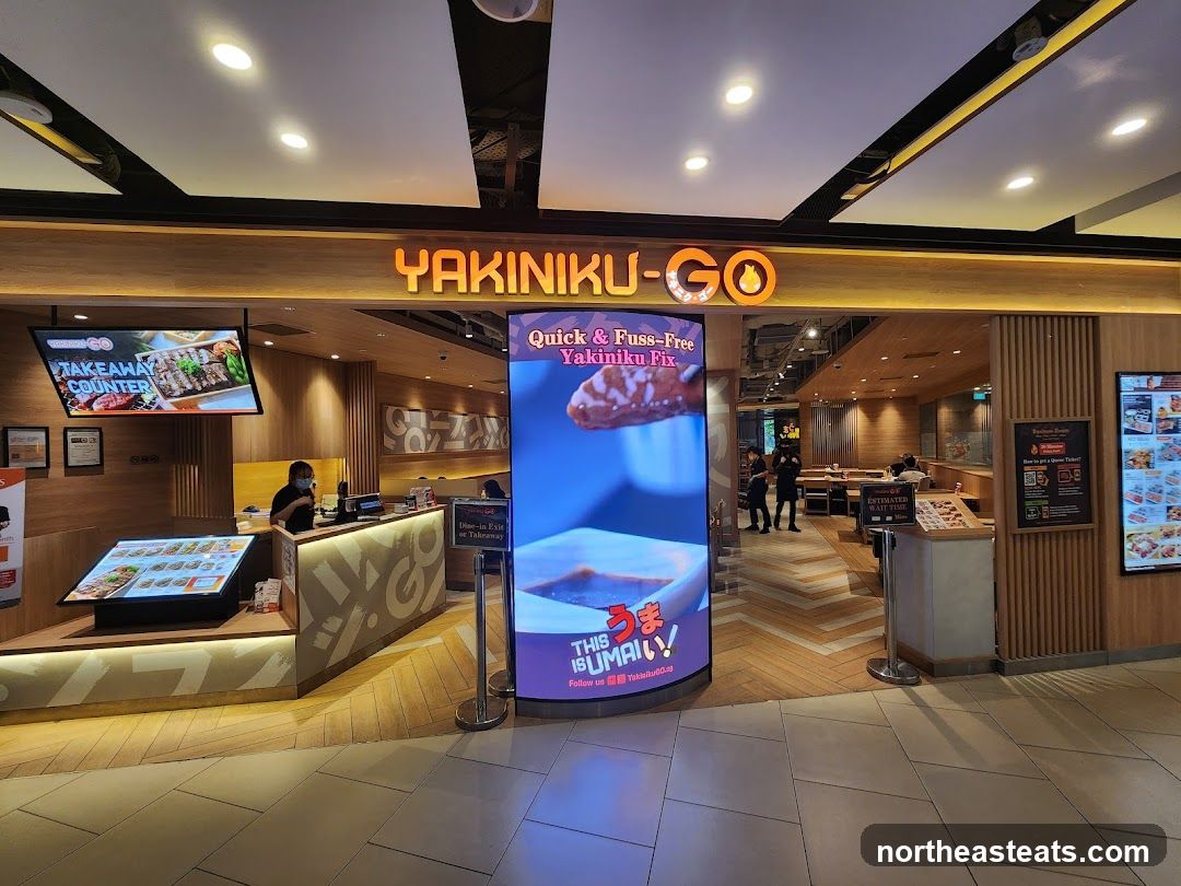 The exterior of a Yakiniku-Go restaurant. A large illuminated sign above the entrance reads "YAKINIKU-GO". To the left, a digital screen displays "TAKEAWAY COUNTER" with images of food. To the right, a large curved screen shows a close-up of food with Japanese text. Inside, staff can be seen behind a counter, and dining tables are visible. A sign indicates "ESTIMATED WAIT TIME".