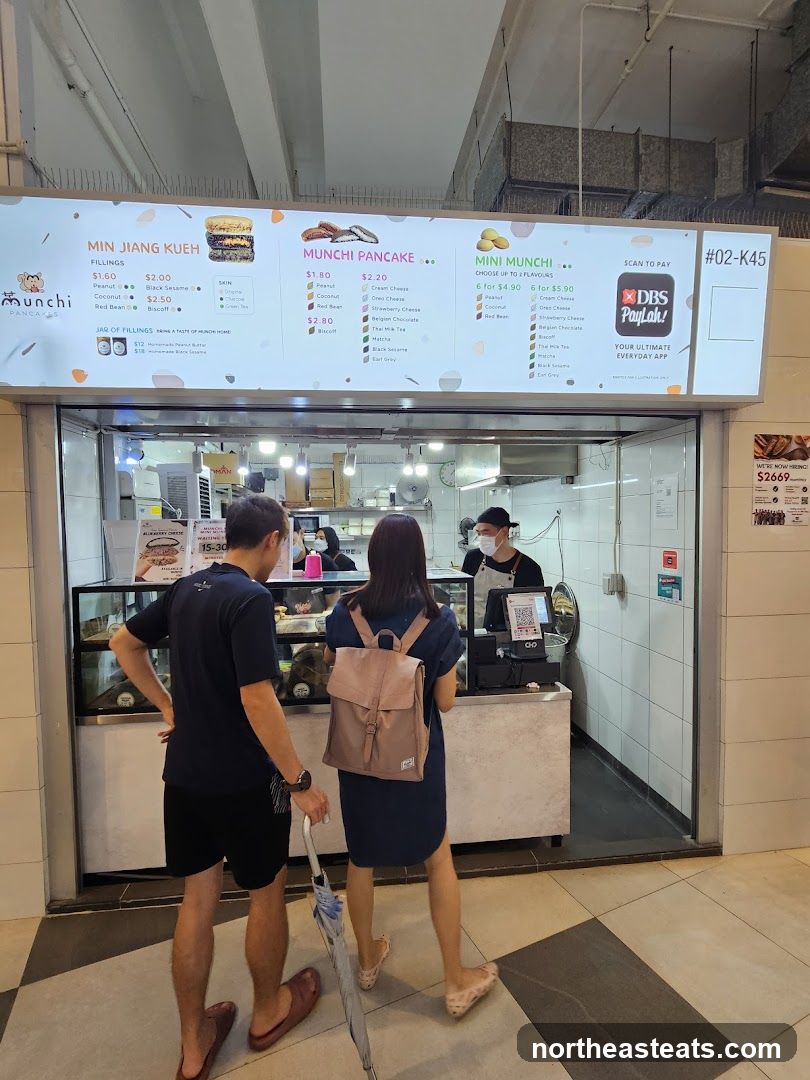 A food stall with a bright overhead menu displaying various pancake and kueh options. Two customers are standing in front of the counter, waiting to order. Staff members are visible behind the counter preparing food.