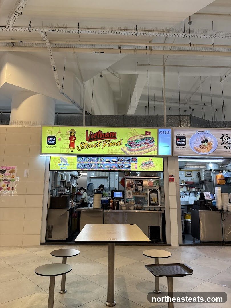 A food stall with a large yellow sign displaying "Vietnam Street Food" with an illustration of a banh mi sandwich. Below the main sign is a menu board with various food items and prices. The stall has a counter with staff preparing food. In front of the stall, there are tables and stools for customers.