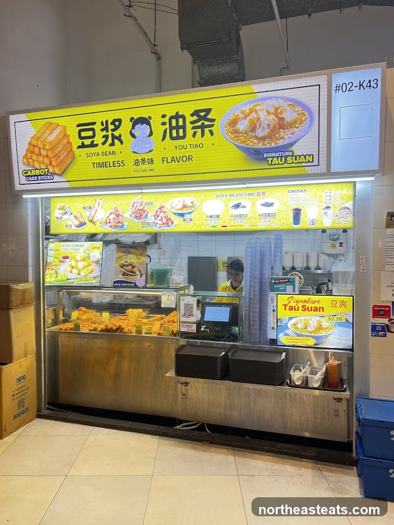 A brightly lit food stall with a large yellow sign featuring the shop's name in Chinese and English, along with images of food. The stall displays various fried items and beancurd in a refrigerated case. A menu board above the case shows pictures and prices of different dishes and drinks. A staff member in a yellow shirt is visible behind the counter.