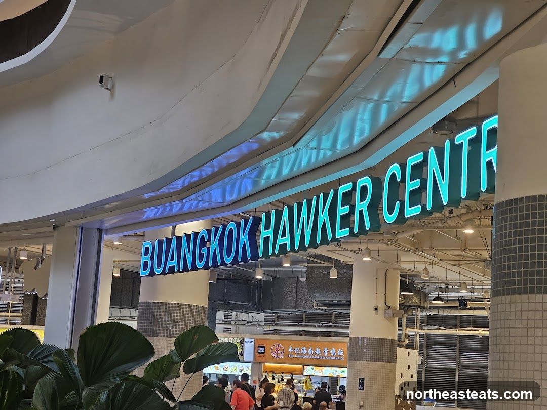 A neon sign that reads "BUANGKOK HAWKER CENTRE" is illuminated in bright blue. The sign is mounted above the entrance to a hawker centre, with a glimpse of stalls and people inside. Large green leaves are in the foreground.
