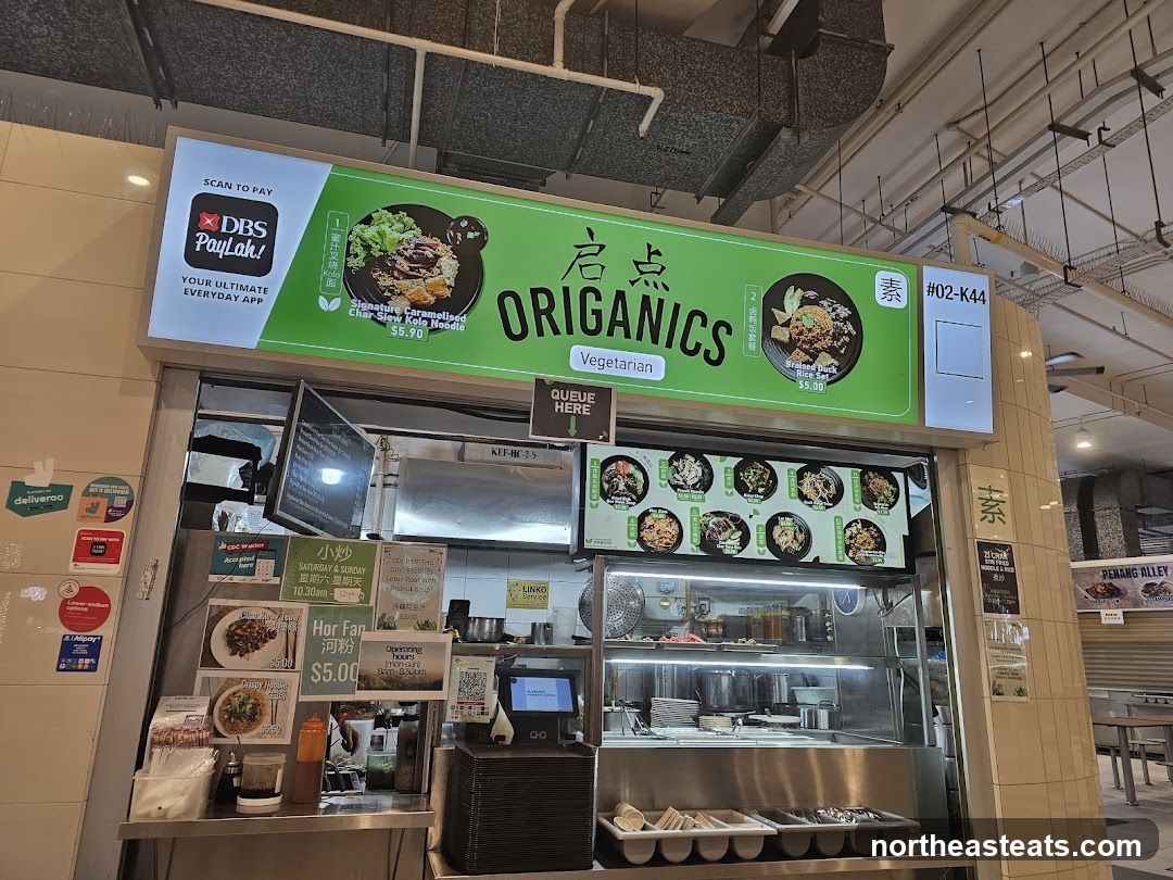A stall with a bright green sign displaying the name "Organics" and "Vegetarian". The sign features images of two dishes: "Signature Caramelised Char-Siew Kolo Noodle" for $5.90 and "Braised Duck Rice" for $5.00. Below the main sign, a digital menu displays various other dishes with images. The stall also has smaller signs advertising "Hor Fan" for $5.00 and operating hours. Delivery service logos like Deliveroo and payment options like DBS PayLah! and Alipay are visible on the left side.