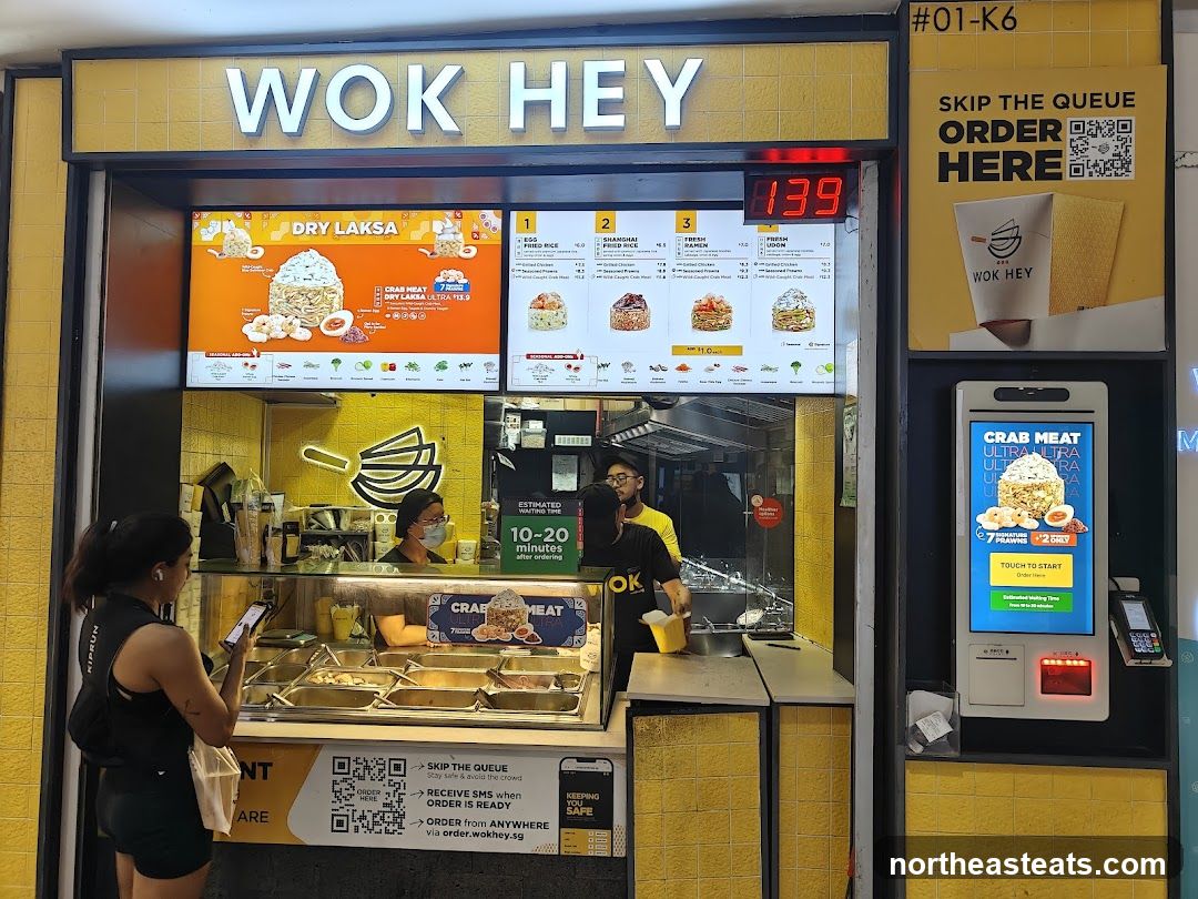 A bright yellow and white food stall with the name "WOK HEY" prominently displayed. A digital menu board shows various noodle dishes, including "DRY LAKSA" and "SHANGHAI FRIED RICE". Customers can order via a QR code or a touch screen kiosk. Staff members are visible behind the counter preparing food.