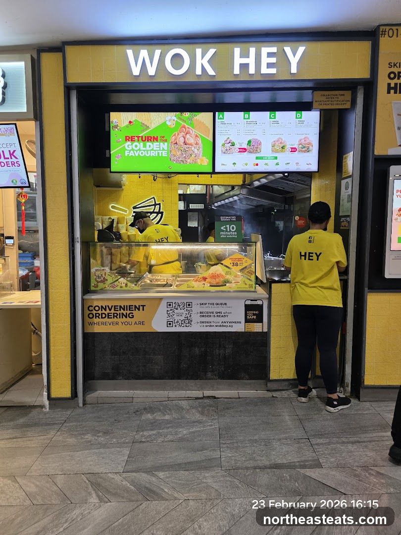 A bright yellow storefront with the name "WOK HEY" displayed in large white letters above the entrance. Digital screens display food images and menu options. A staff member in a yellow shirt with "HEY" on the back is visible inside. A sign promotes convenient ordering via QR code and a website.