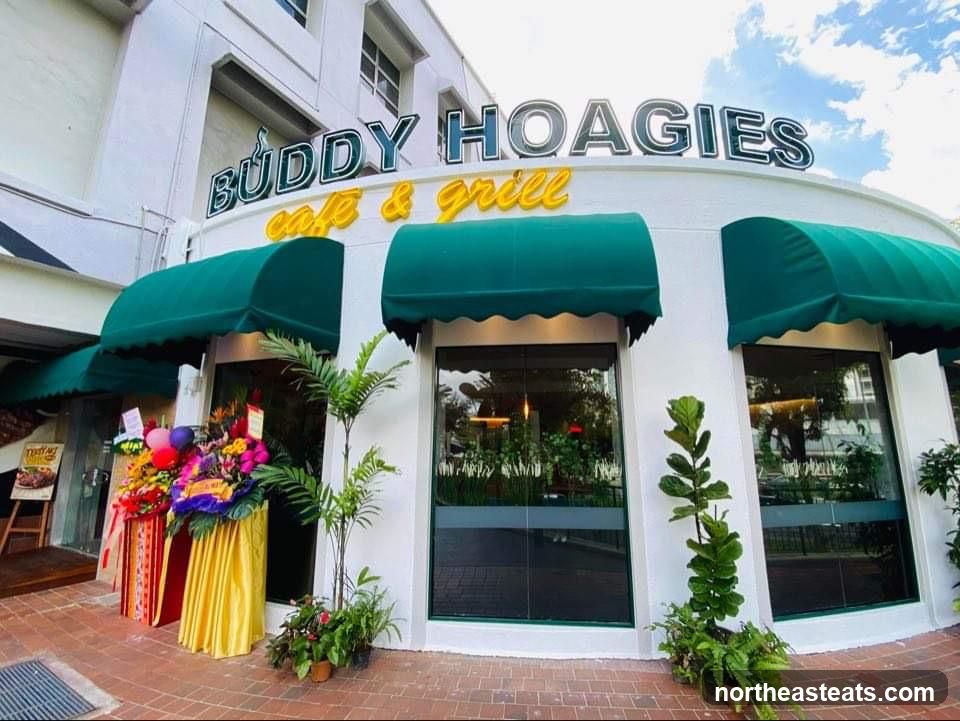 The exterior of a cafe and grill with a white facade, green awnings, and large windows. The business name "BUDDY HOAGIES cafe & grill" is prominently displayed in neon lettering above the entrance. There are floral arrangements and balloons outside the entrance, suggesting a special occasion.