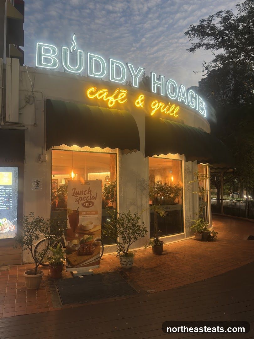 The exterior of Buddy Hoagies Cafe & Grill at dusk. Neon signs spell out the name of the establishment. A green awning covers the windows, and potted plants are placed outside.