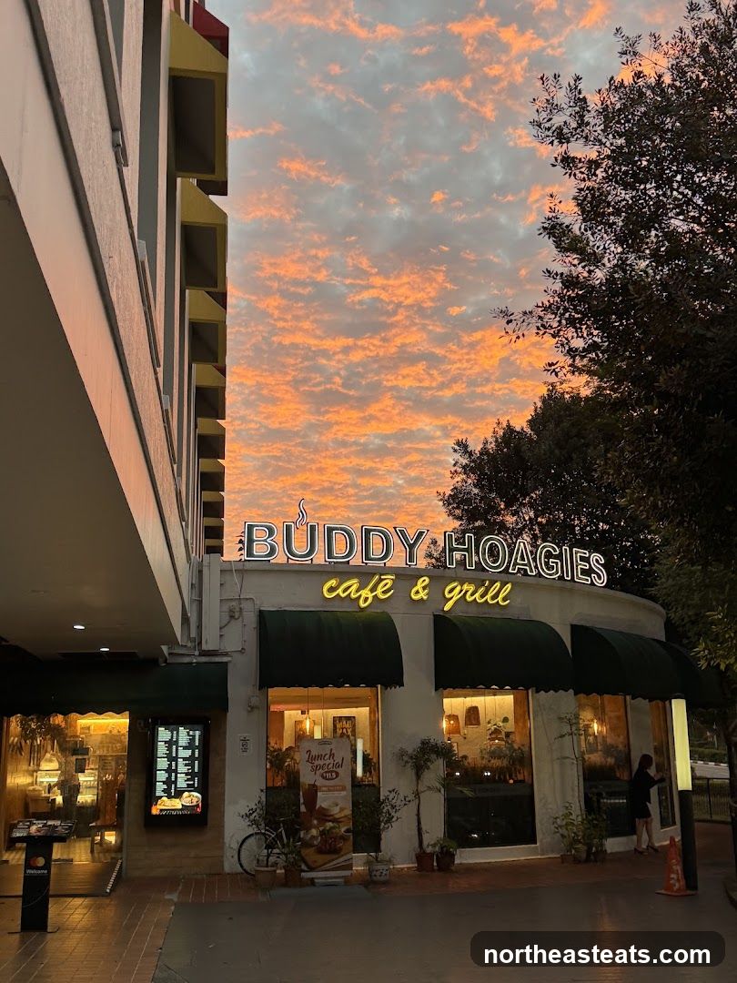 The exterior of Buddy Hoagies Cafe & Grill at sunset. The cafe has a neon sign above the entrance. There are green awnings over the windows. Inside, the cafe is lit and appears to be open. A lunch special sign is visible outside.